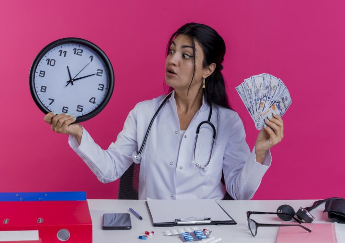impressed young female doctor wearing medical robe and stethoscope sitting at desk with medical tools holding money and clock looking at clock isolated on pink background independência financeira materna